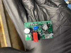 Na zdjęciu znajduje się mała płytka elektroniczna trzymana w ręce w czarnej rękawiczce. Płytka ma kilka komponentów, takich jak kondensatory i inne elementy elektroniczne, oraz oznaczenie MH-ACG2 22.04.18.