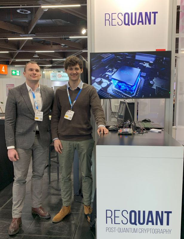 Stoisko firmy ResQuant na targach, dwóch mężczyzn pozuje obok monitora pokazującego układ scalony.