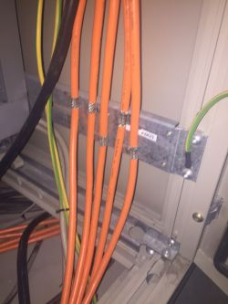 Pomarańczowe kable i inne przewody w środku urządzenia elektrycznego, zamocowane na metalowym uchwycie.
