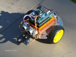 A robot built with ESP32 and UNO boards, mounted on a metal chassis with yellow wheels and an ultrasonic sensor in front.