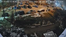 Widok z kamery monitoringu na osiedlowy parking i chodnik zimą nocą, z lekko ośnieżonymi samochodami.