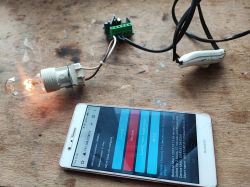 A smartphone displaying an online control panel next to a lit bulb connected by wires to an electronic setup on a wooden table.