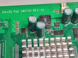 Zbliżenie na fragment płytki drukowanej switcha 24+2G PoE, widoczne kondensatory elektrolityczne, cewka i radiator aluminiowy.