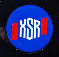 Okrągły ekran z niebieskim tłem i białym napisem „XSR” oraz czerwonymi blokami po bokach
