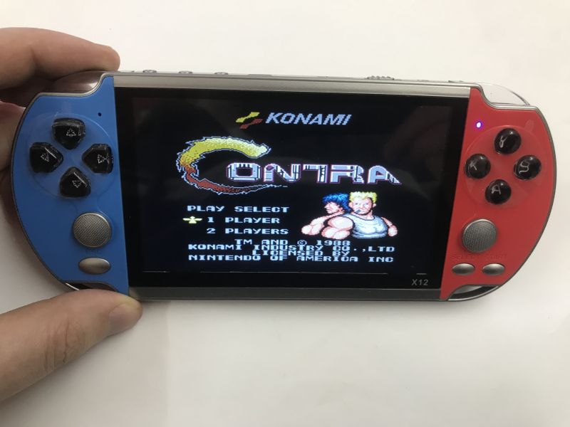 X12 handheld console held in a hand, showing the “Contra” title menu with the Konami logo.