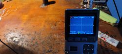 Compact digital oscilloscope on a workbench displaying a waveform on the screen