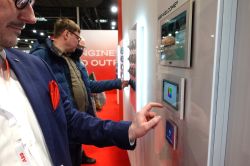 Person touches an ABB touchscreen panel at a trade show booth; other visitors and red carpet in the background.