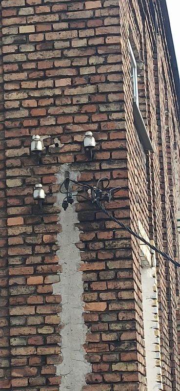 Ceglana ściana starej kamienicy z porcelanowymi izolatorami i widocznymi przewodami elektrycznymi.