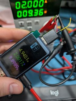 Dłoń trzyma tester USB-C z ekranem pokazującym napięcie, prąd i moc; w tle zasilacz z zielonym wyświetlaczem