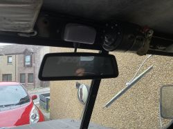 Lusterko w kabinie pojazdu z lampką sufitową; przez szybę widać wycieraczkę i czerwone auto na zewnątrz