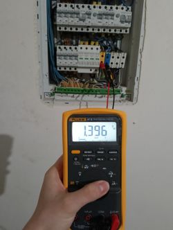 Wyświetlacz multimetru pokazujący napięcie 1,396 V AC przed rozdzielnicą elektryczną.