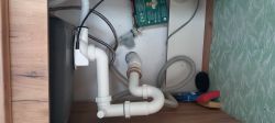 Widok wnętrza szafki pod zlewem z instalacją hydrauliczną i akcesoriami do sprzątania.