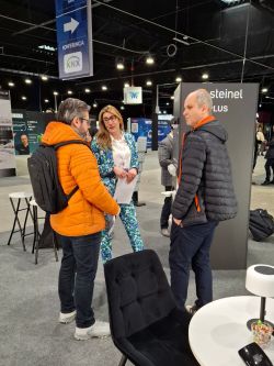 Three people talk at an exhibition booth; “steinel” sign and “KONFERENCJA KNX” direction sign visible.
