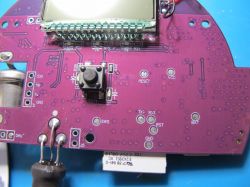 Close-up of a purple printed circuit board with an LCD display and a test button.
