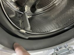 Zbliżenie na wnętrze bębna pralki z widocznym plastikowym elementem wystającym nad bębnem oraz palcem wskazującym gumową uszczelkę.