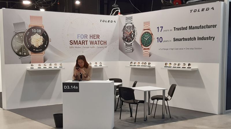 Stoisko z inteligentnymi zegarkami marki Toleda na targach elektroniki, z siedzącą osobą obsługującą telefon.