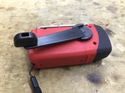 Red and black emergency radio with a hand crank dynamo lying on a wooden surface.