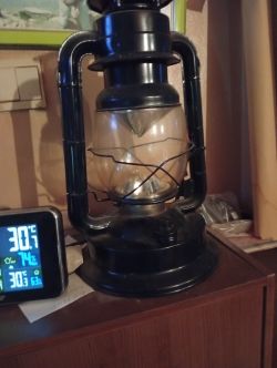 Czarna lampa naftowa o klasycznym wyglądzie stoi obok cyfrowego termometru.
