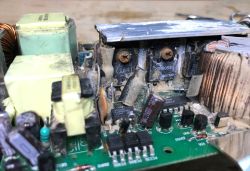 Zbliżenie na uszkodzony i skorodowany zasilacz ze starego switcha, widoczne elementy elektroniczne i ślady zabrudzeń.