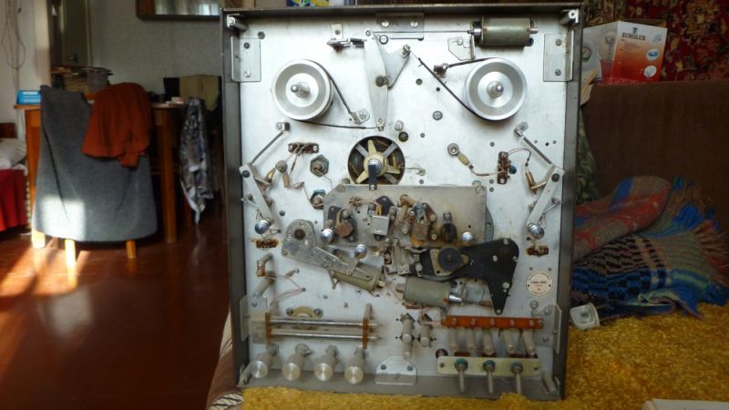 Open reel-to-reel tape recorder mechanism with pulleys and levers, placed in a room