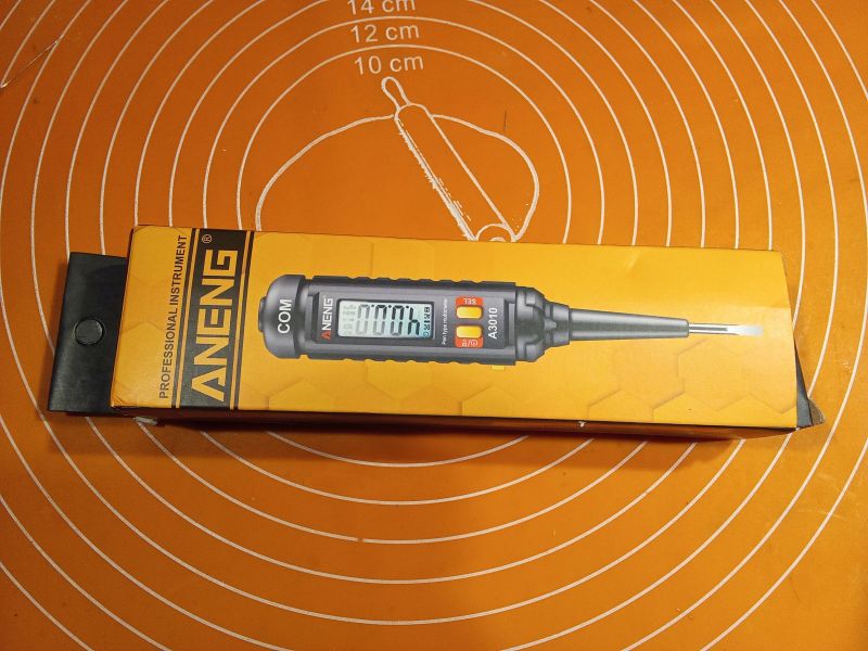 Pudełko z wielofunkcyjnym miernikiem ANENG leżące na pomarańczowej macie kuchennej.