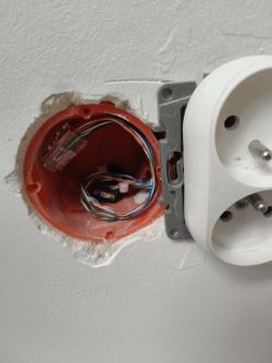 Otwarta puszka instalacyjna w ścianie z widocznymi przewodami sieciowymi obok gniazdka elektrycznego.