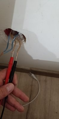 Zbliżenie na ścianę z wyprowadzonymi przewodami elektrycznymi i sondami multimetru trzymanymi przez rękę.