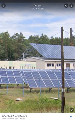 Elektrownia słoneczna z panelami fotowoltaicznymi na tle budynku i lasu.