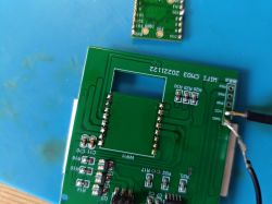 PCB with a removed WiFi module, visible solder pads and pin rows, on a blue work surface.