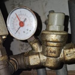 Reduktor ciśnienia na rurze z manometrem; wskazówka ok. 5 bar, metalowy korpus i białe pokrętło.