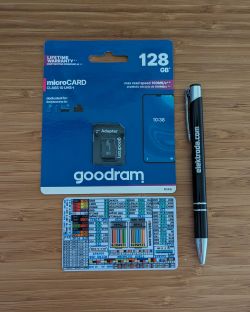 Goodram 128GB microSD card in packaging, lying on a wooden surface next to a pen and a colorful card with diagrams.