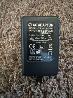 Schwarzes Netzteil mit 12V DC 1,5A Ausgang auf braunem Teppich