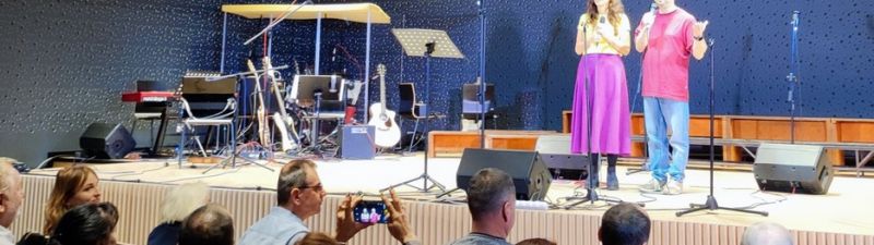 Dwie osoby na scenie stojące przed mikrofonami, z widownią siedzącą przed nimi i sprzętem muzycznym w tle.