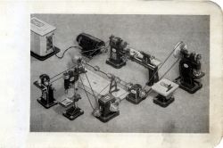 Czarno-białe zdjęcie przedstawiające miniatury maszyn ślusarskich i stolarskich połączonych za pomocą wałów transmisyjnych.