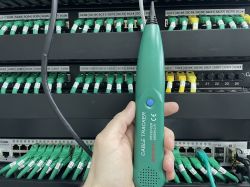 Ręka trzymająca detektor kabli przed panelem krosowniczym z podłączonymi kablami ethernetowymi.