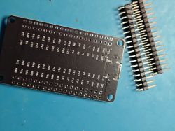 Black PCB with labeled pin holes, next to two pin header strips on a blue mat