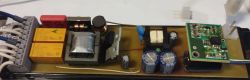 Otwarta płytka elektroniki z kondensatorami, transformatorami i dwoma żółtymi przekaźnikami przy listwie zaciskowej
