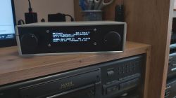 A modern DIY internet radio with an LCD display on a wooden shelf, showing track information.