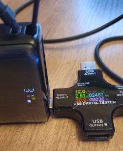 Powerbank podłączony do miernika USB z ekranem pokazującym 12,0 V, 2,51 A i 30,2 W na biurku