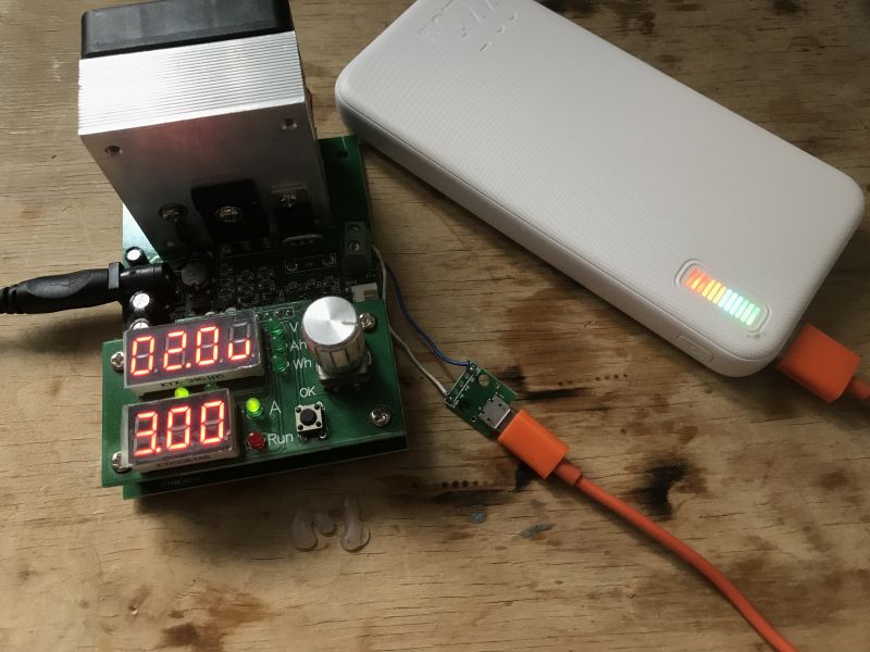 Powerbank podłączony do urządzenia testującego z wyświetlaczami pokazującymi napięcie i natężenie.