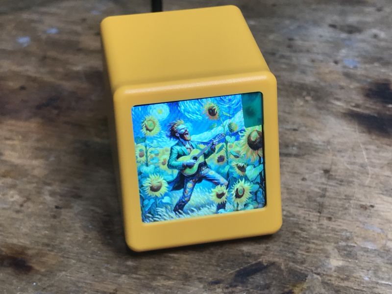 Gelbes SmallTV-Ultra-Gerät mit Bild auf dem Display Gelbes SmallTV-Ultra-Gerät mit einem Display, das ein Bild eines Künstlers, umgeben von Sonnenblumen, zeigt.