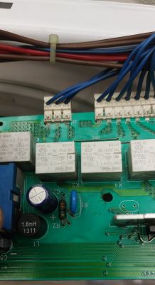 Zbliżenie na płytkę elektroniczną z różnymi komponentami, takimi jak przekaźniki i kondensatory, w tym niebieski element.