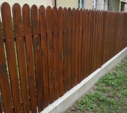 Drewniany płot z pionowych sztachet, pomalowany na brązowo, na betonowej podmurówce