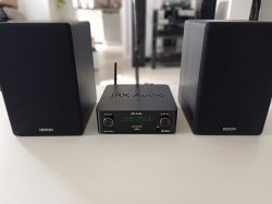 Amplituner JRK Audio Evothon ustawiony między dwoma głośnikami Denon SC-N10 na białym blacie.