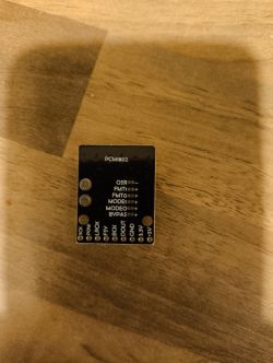 Black PCM1802 ADC module on a wooden surface, with printed pin labels and configuration jumpers.