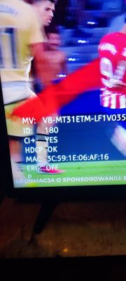 Ekran telewizora w trybie serwisowym z informacjami technicznymi i rozmazanym obrazem meczu piłki nożnej w tle.