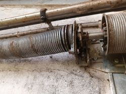 Widok mechanizmu sprężynowego bramy garażowej z widoczną sprężyną, zębatką i rdzą na częściach.