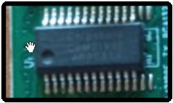 Zbliżenie na układ scalony SMD z wieloma wyprowadzeniami na zielonej płytce PCB; napisy są nieczytelne.