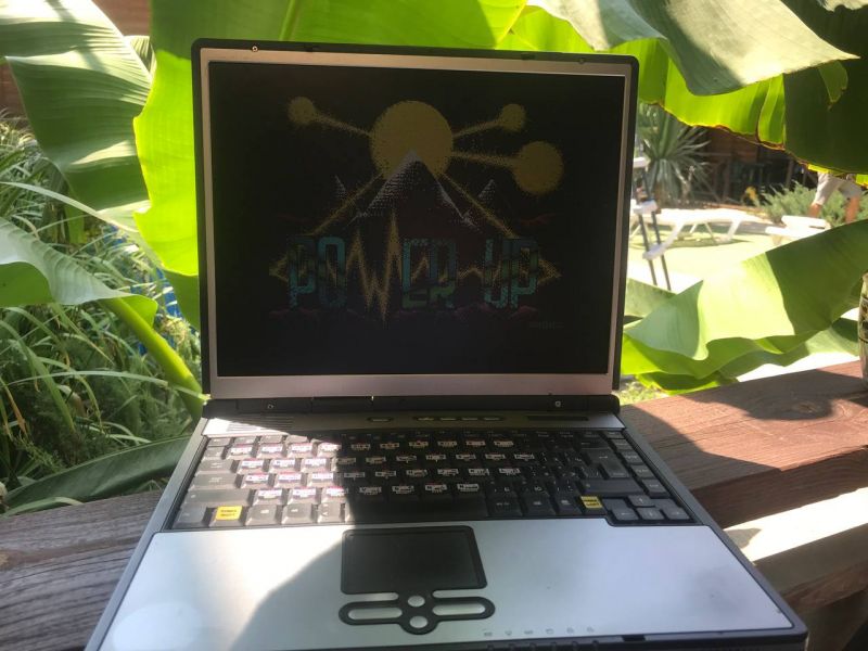 Laptop z napisem Power Up na ekranie, stojący na tle roślin.