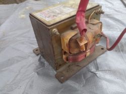Transformator w metalowej obudowie, z widocznymi zwojami miedzianego drutu i złączami elektrycznymi.